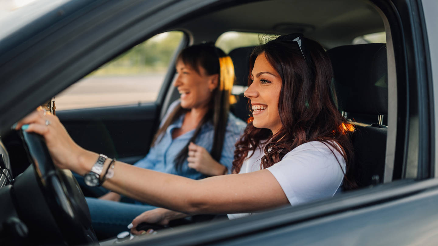 Smiling fashionable female friends sitting in a car and driving it on city road.Side view of trendy girl driving car with hands on steering wheel while her best friend is sitting next to her, laughing