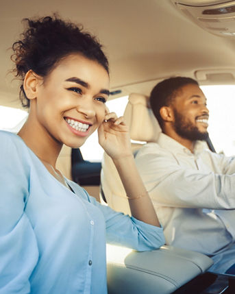 African American happy couple enjoys a car ride on a sunny day. The woman, with a big smile on her face, is in the passenger seat, while the man, wearing a white shirt, is driving.