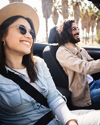 Young woman smiling at camera while sitting in convertible car. Man driving while couple enjoys sunny summer journey together. Travel and lifestyle concept.