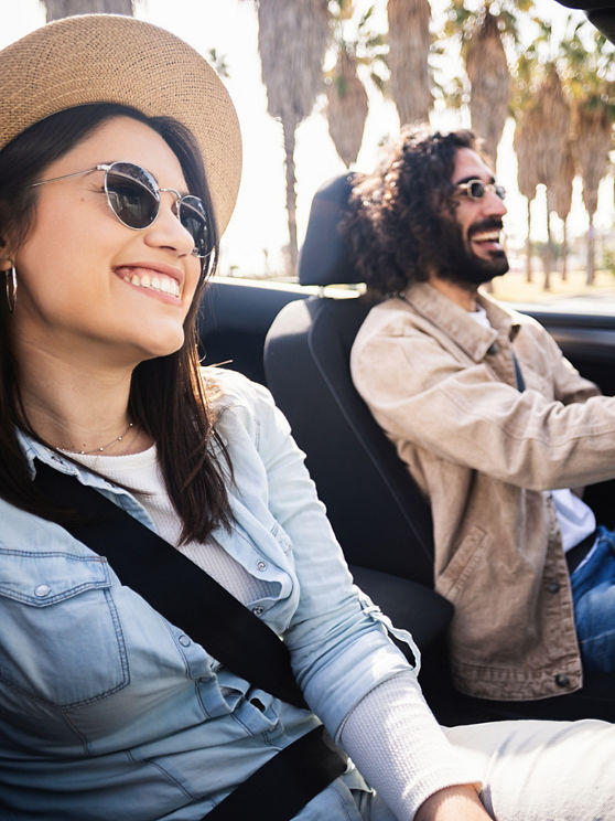 Young woman smiling at camera while sitting in convertible car. Man driving while couple enjoys sunny summer journey together. Travel and lifestyle concept.