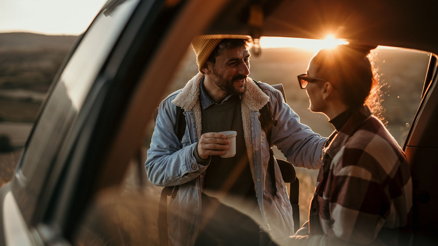 Young couple talking, drinking a coffee watching the sunset from their car.